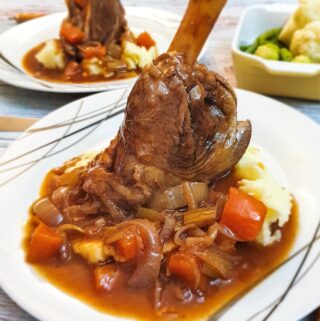 A lamb shank on a pile of mashed potatoes covered with red wine gravy and vegetables.
