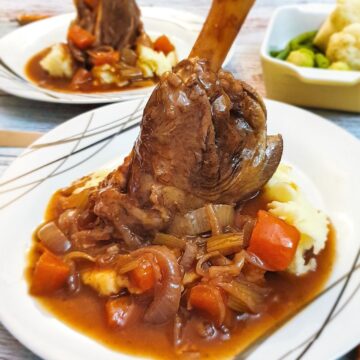 A lamb shank on a pile of mashed potatoes covered with red wine gravy and vegetables.