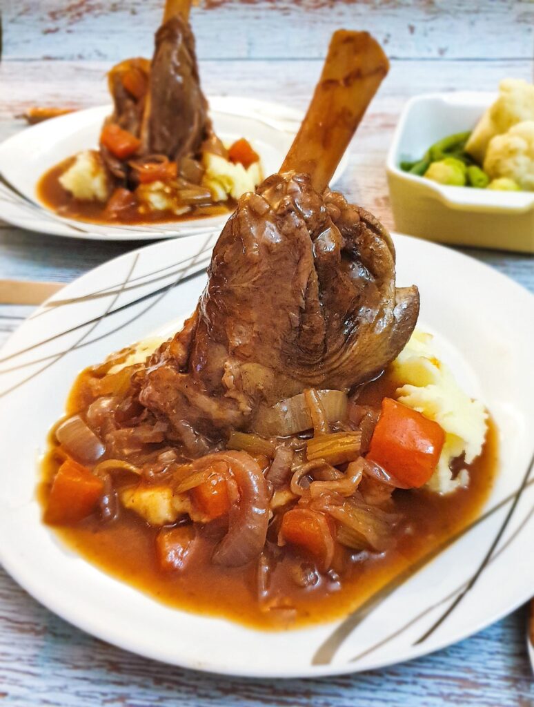 A lamb shank on a pile of mashed potatoes covered with red wine gravy and vegetables.