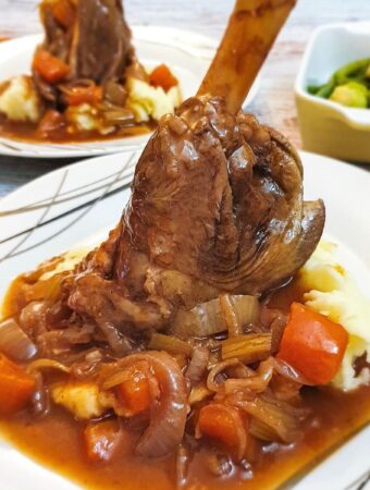 A lamb shank on a pile of mashed potatoes covered with red wine gravy and vegetables.