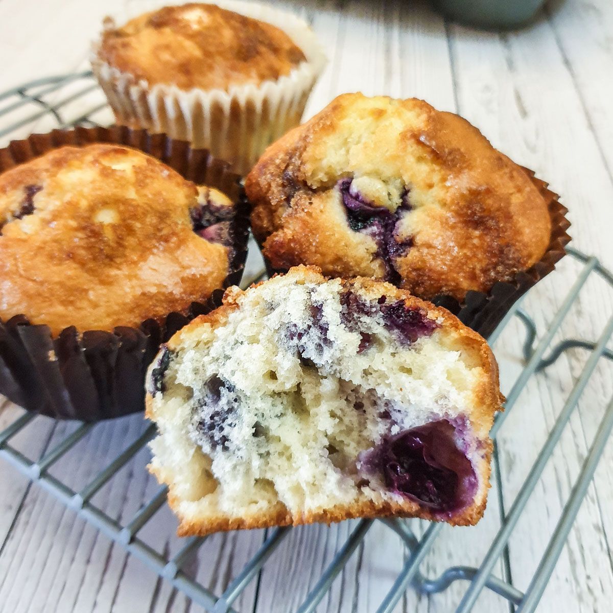 Half a blueberry muffin showing the texture on a cooling rack in front of three blueberry muffins.