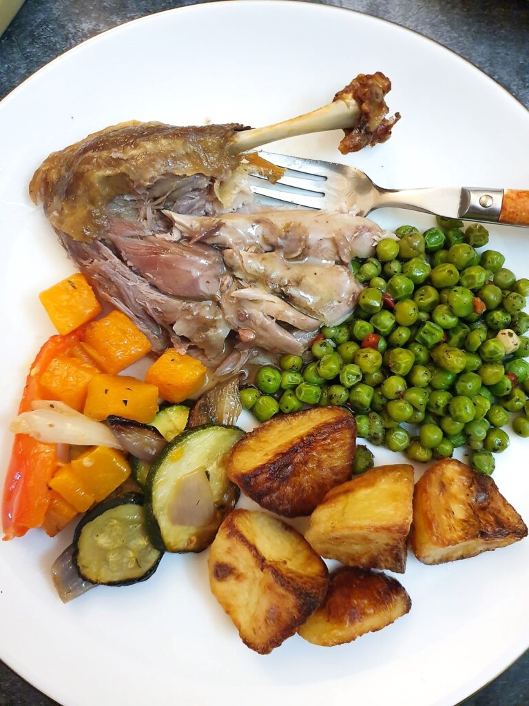 A cooked duck leg on a plate with potatoes and vegetables.