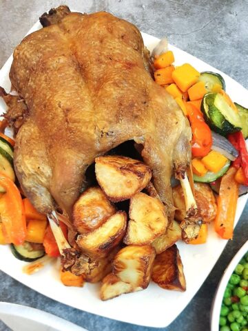 A roast duck on a serving plate with roast potatoes and vegetables.