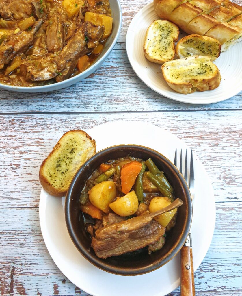 A dish of lamb bredie on a table with a slice of garlic bread on the side.