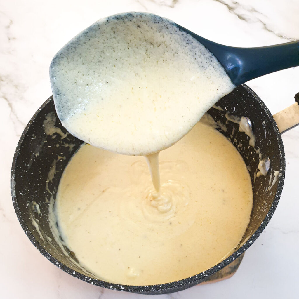 Cheese sauce being poured from a large spoon into a saucepan, showing the consistency.