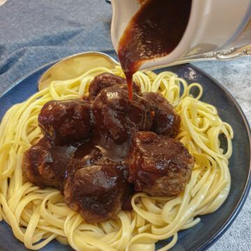 A jug of barbeque sauce being poured over a dish of linguine and meatballs.