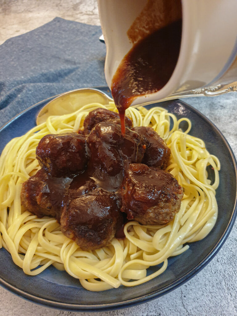 A jug of barbeque sauce being poured over a dish of linguine and meatballs.