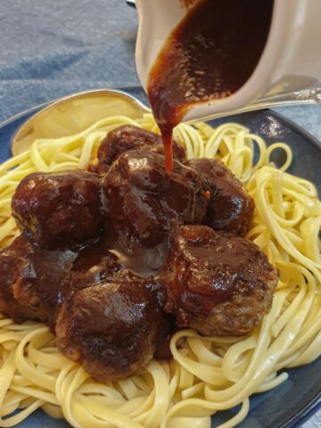 A jug of barbeque sauce being poured over a dish of linguine and meatballs.