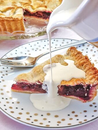 A jug of cream being poured over a slice of blackberry and apple pie.