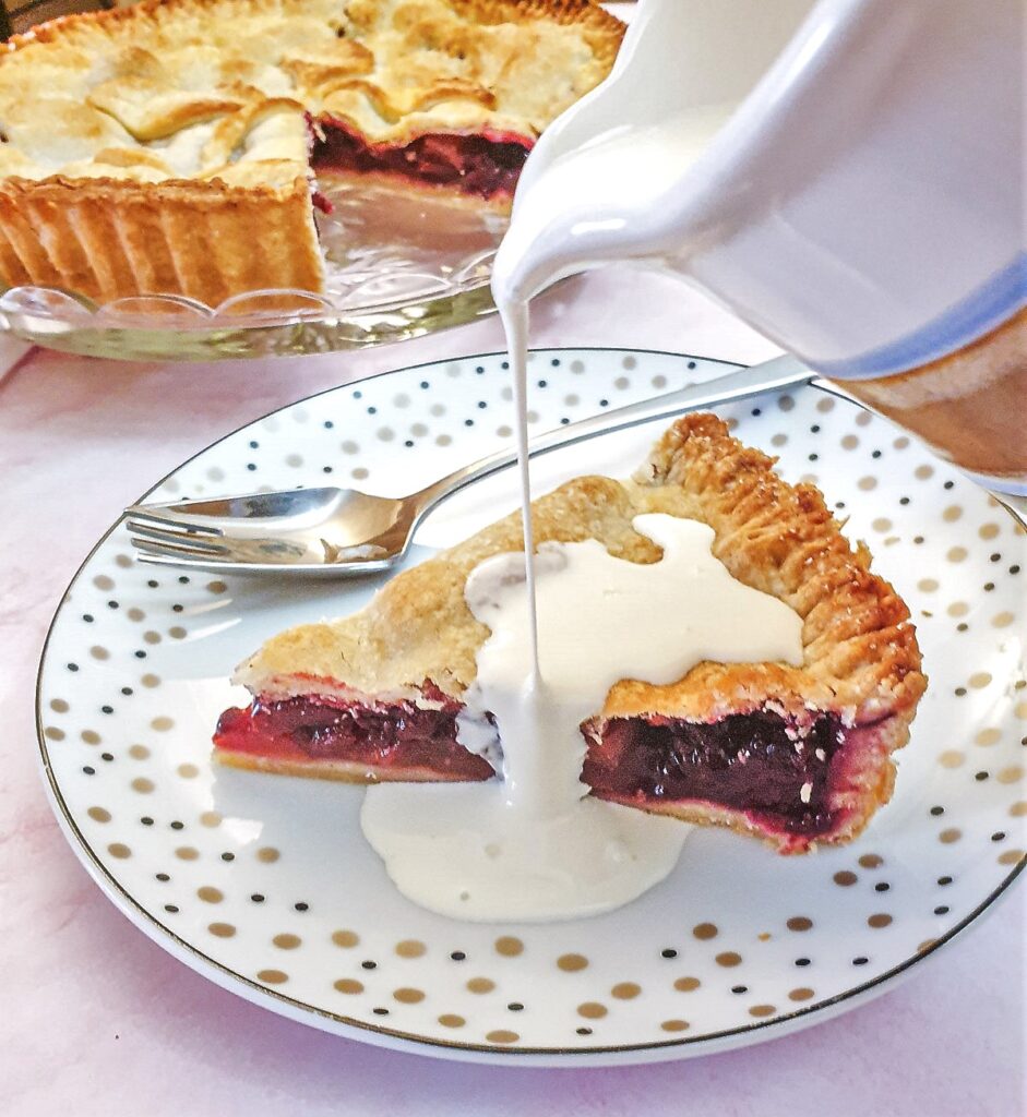 A jug of cream being poured over a slice of blackberry and apple pie.