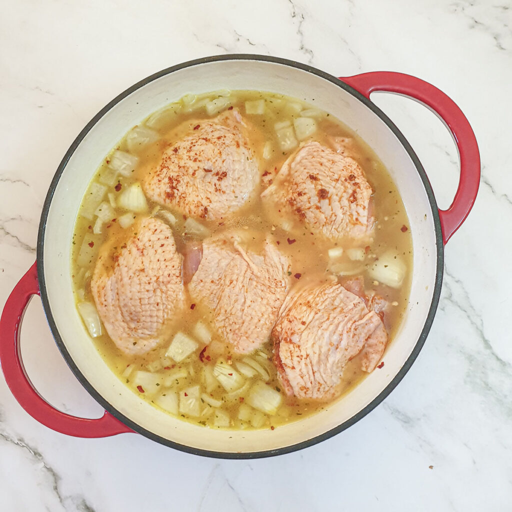 Chicken pieces on top of a pan of chicken stock with rice and onions.