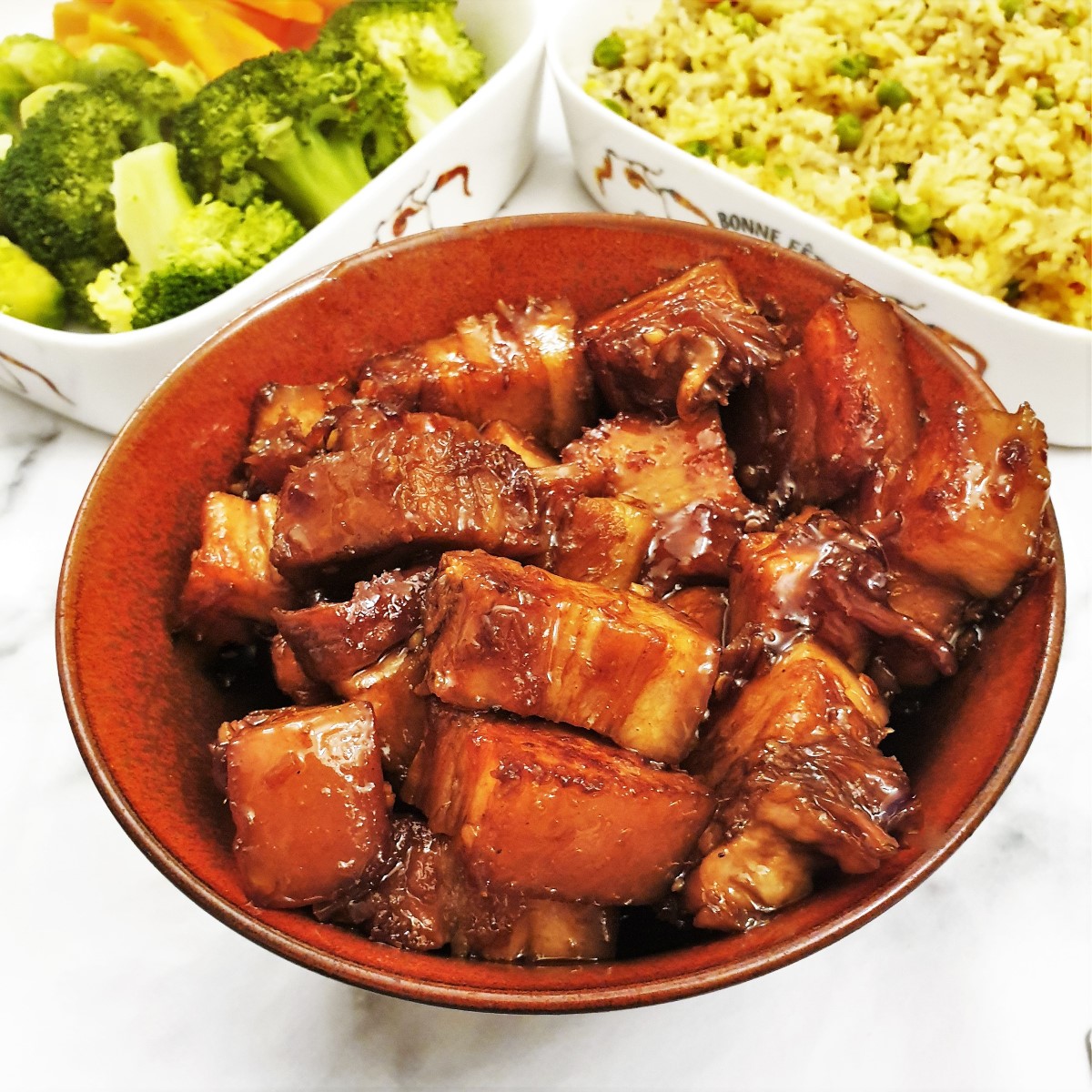 A dish of sticky pork belly next to a dish of rice and a dish of vegetables.