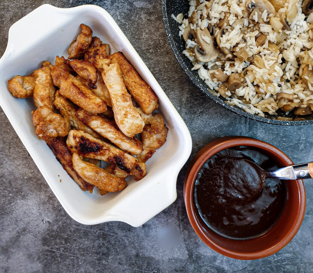 A dish of cooked pork fingers next to a dish of barbeque sauce.