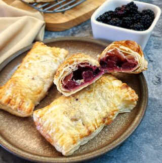 Three blackberry and apple turnovers on a plate with one of them cut in half to show the inside.