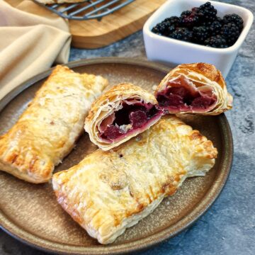 Three blackberry and apple turnovers on a plate with one of them cut in half to show the inside.