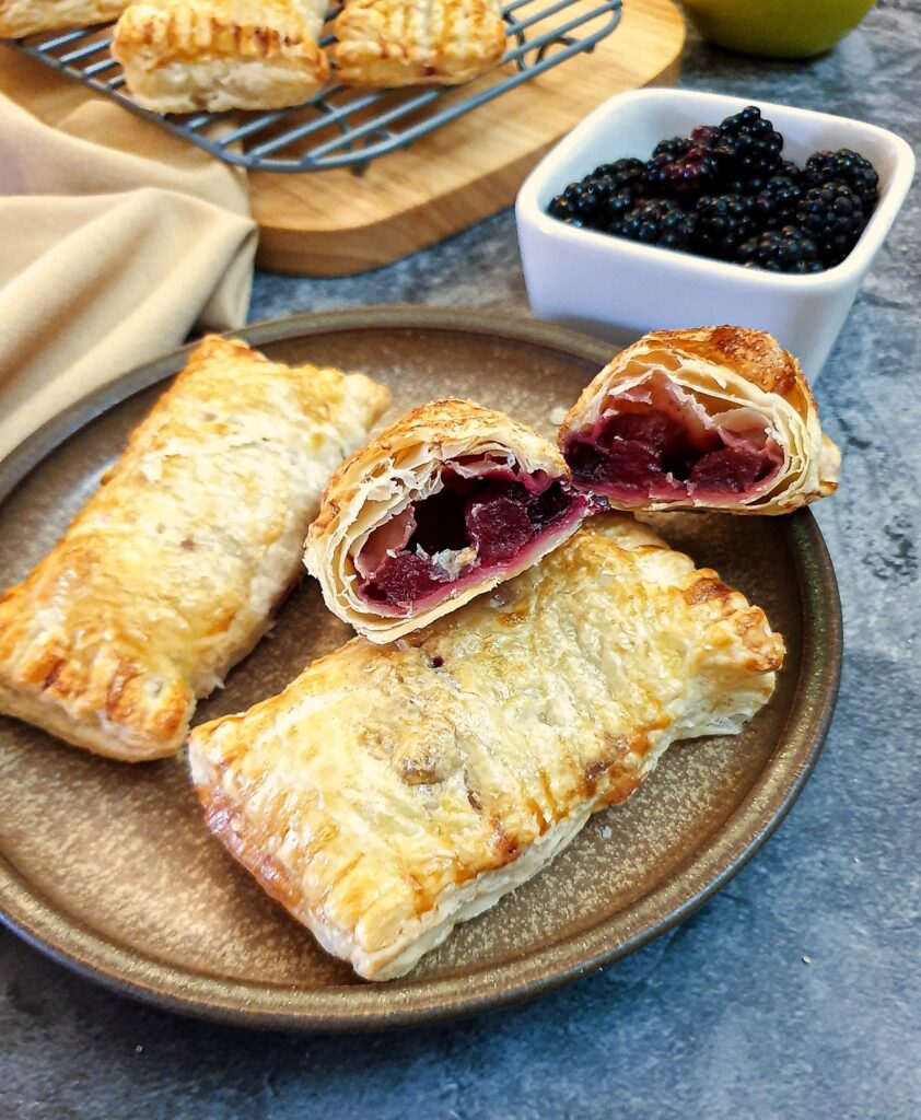 Three blackberry and apple turnovers on a plate with one of them cut in half to show the inside.