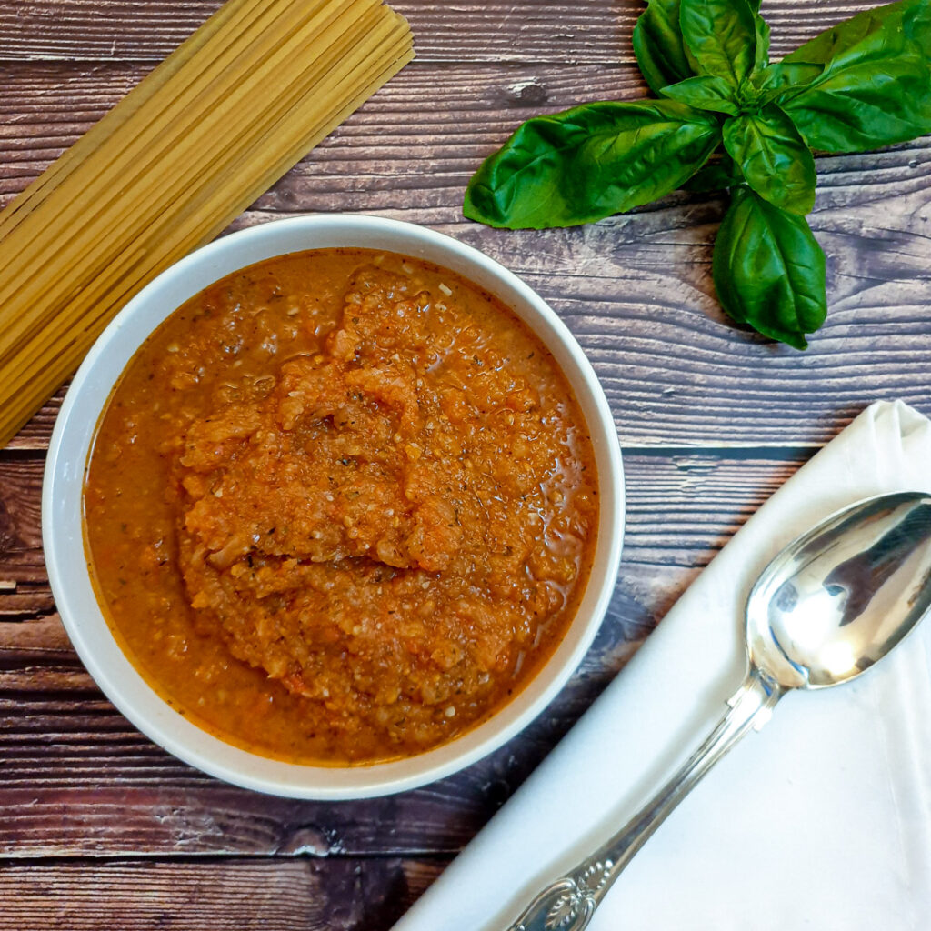 A dish of marinara sauce on a table next to a pile of uncooked spaghetti.