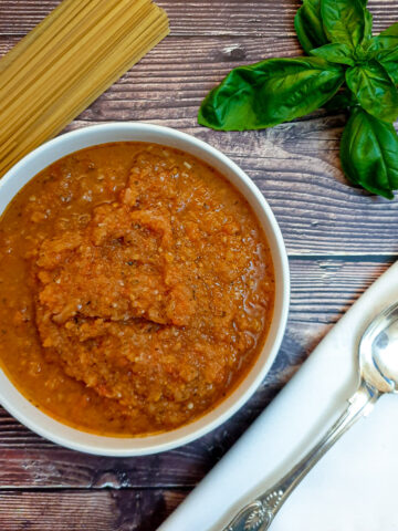 A dish of marinara sauce on a table next to a pile of uncooked spaghetti.