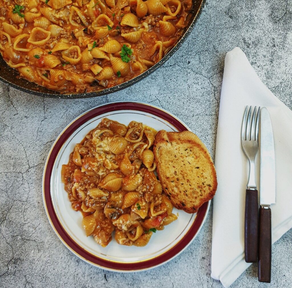 A plate of cheesy mince pasta with a slice on garlic bread on a table.