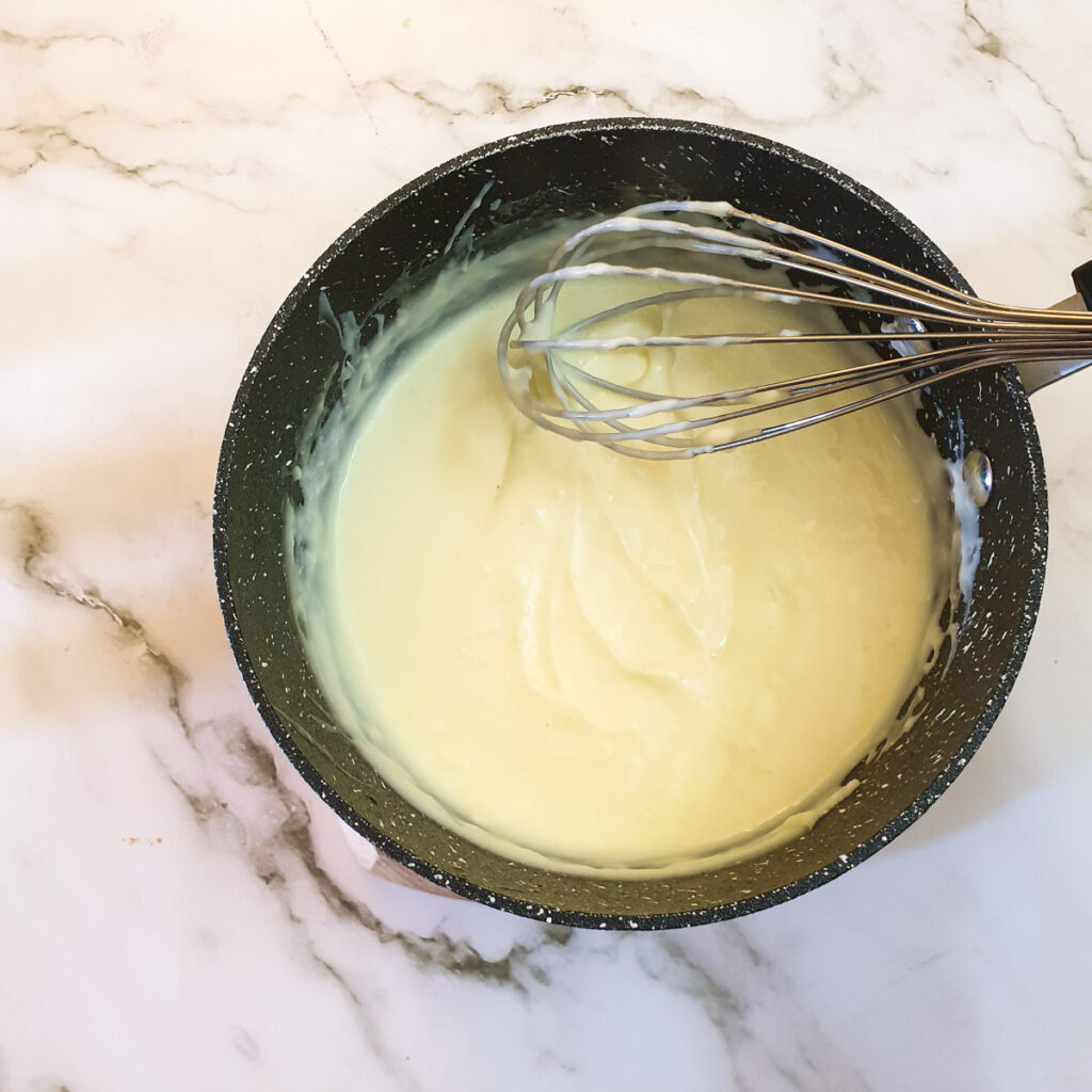 Cheese sauce being stirred with a balloon whisk in a saucepan.