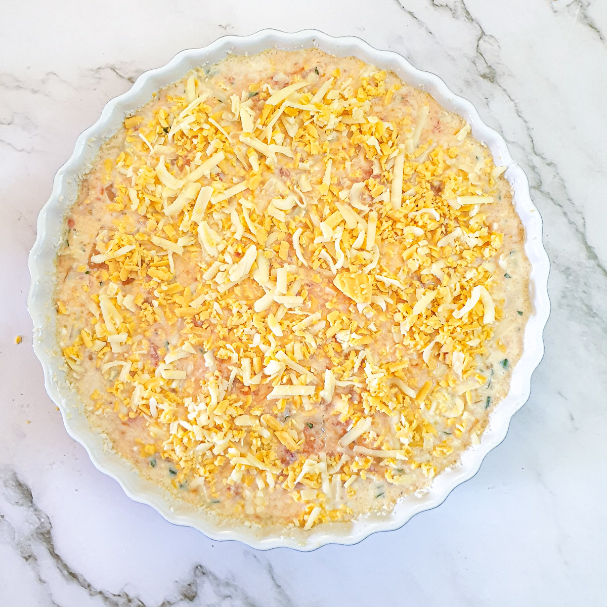 Filling for crustless tomato quiche in a baking dish sprinkled with grated cheese.