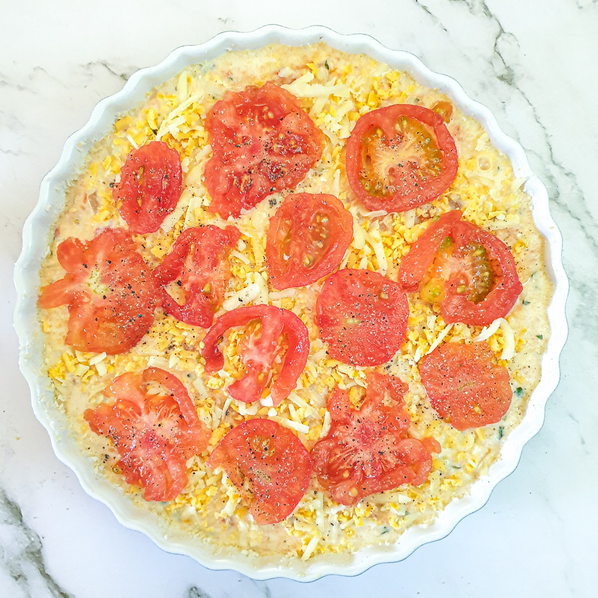 Filling for crustless tomato quiche in a baking dish sprinkled with grated cheese and topped with sliced tomatoes.