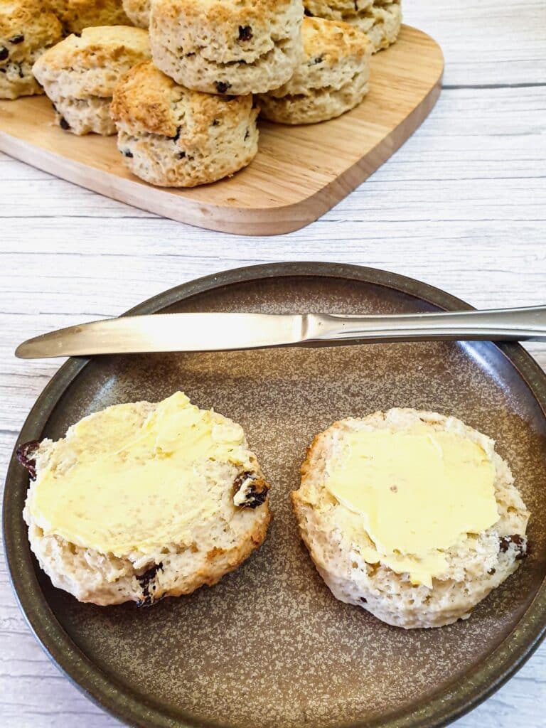 Two buttered sultana scones on a brown plate with a knife.