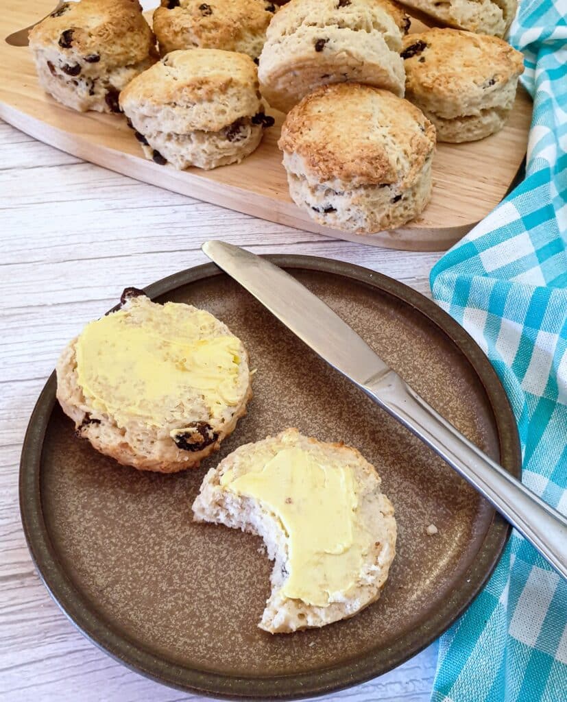 Two halves of buttered sultana scones on a plate with a bite taken out of one.
