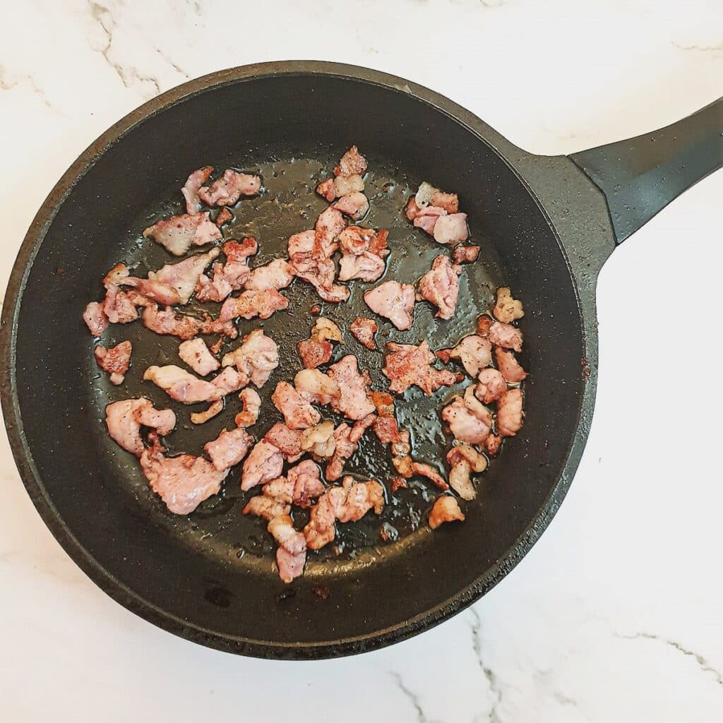 Pieces of bacon being browned in a frying pan.