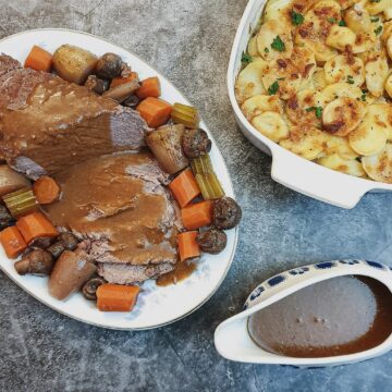 Slices of boeuf bourguignon on a serving dish, surrounded by vegetables, alongside a dish of potatoes and a jug of gravy.