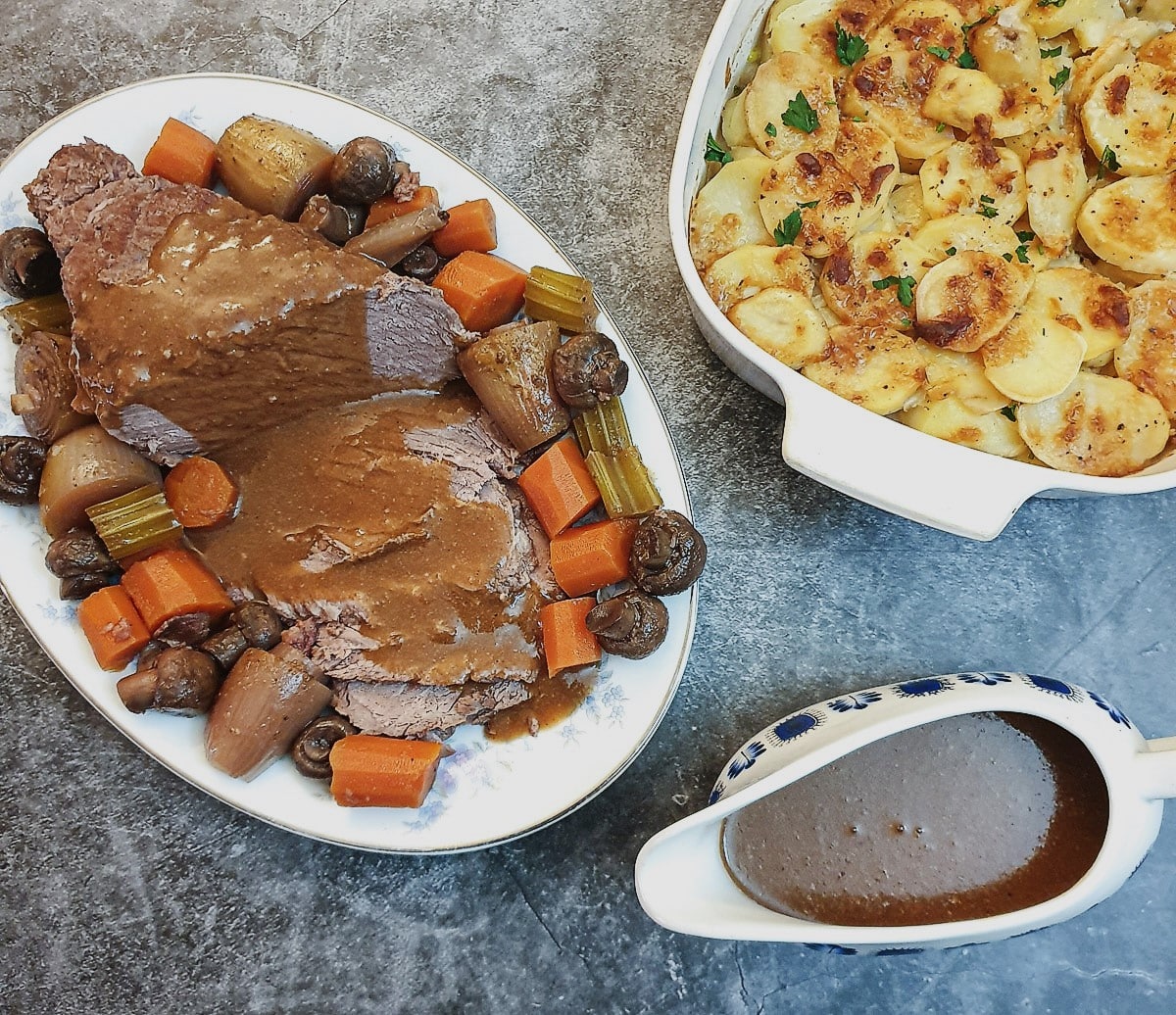 Slices of boeuf bourguignon on a serving dish, surrounded by vegetables, alongside a dish of potatoes and a jug of gravy.