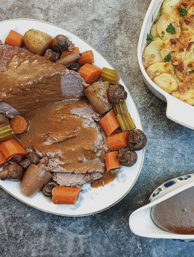 Slices of boeuf bourguignon on a serving dish, surrounded by vegetables, alongside a dish of potatoes and a jug of gravy.
