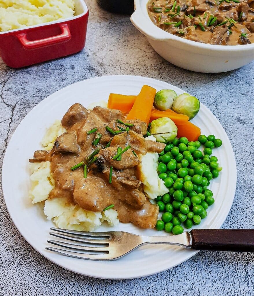 A plate of beef stroganoff on mashed potables with peas and carrots.