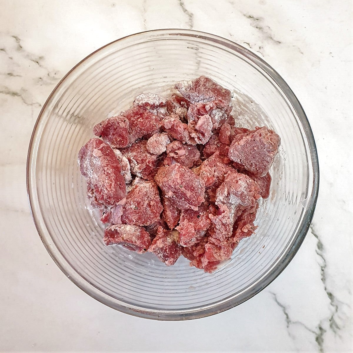 Cubed steak coated with seasoned flour in a glass bowl.