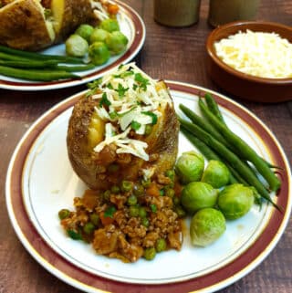 Two baked potatoes covered with savoury mince and grated cheese, with a bowl of grated cheese in the background.