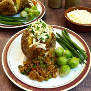 Two baked potatoes covered with savoury mince and grated cheese, with a bowl of grated cheese in the background.