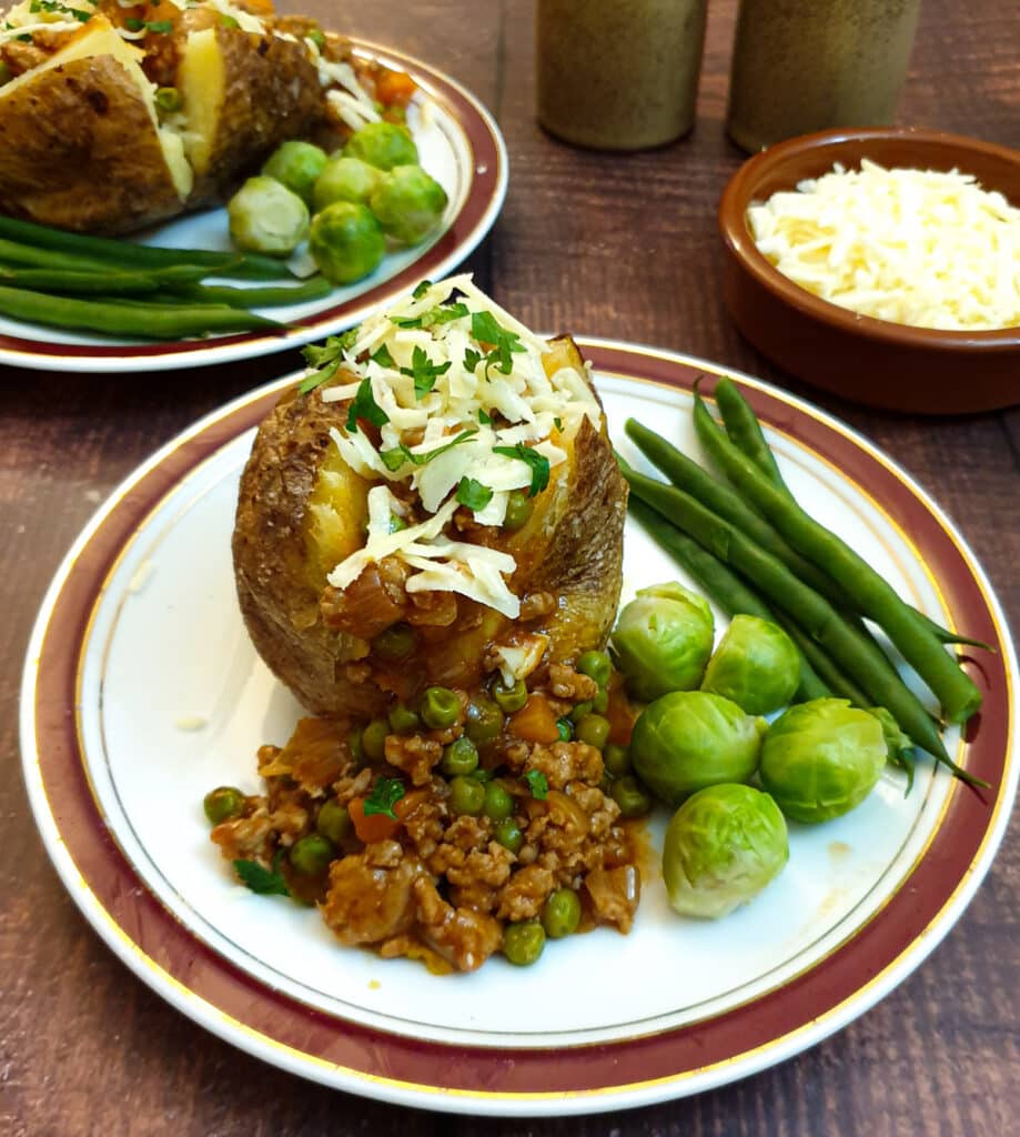 Two baked potatoes covered with savoury mince and grated cheese, with a bowl of grated cheese in the background.