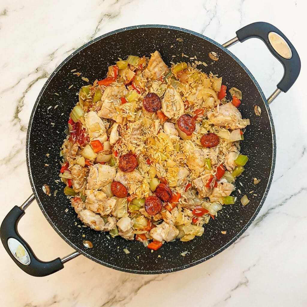 Uncooked rice added to the jambalaya ingredients in a pan.