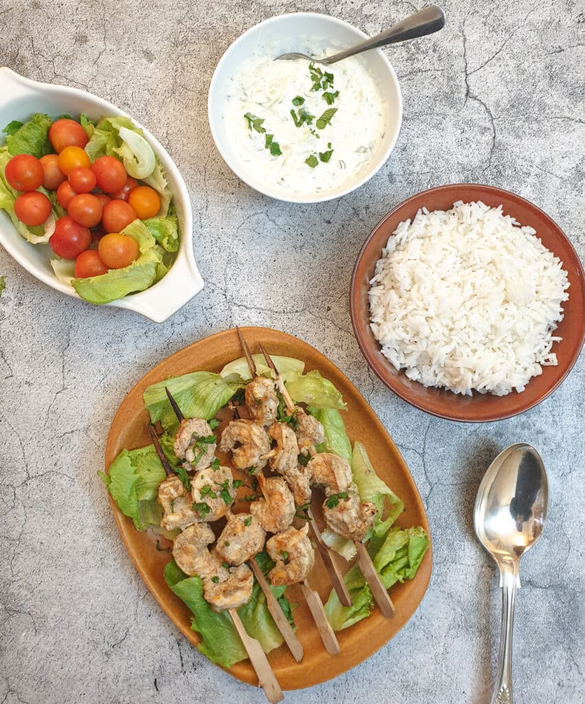 A plate of curried prawn skewers with bowls of rice, tomatoes and cucumber raita, seen from above.