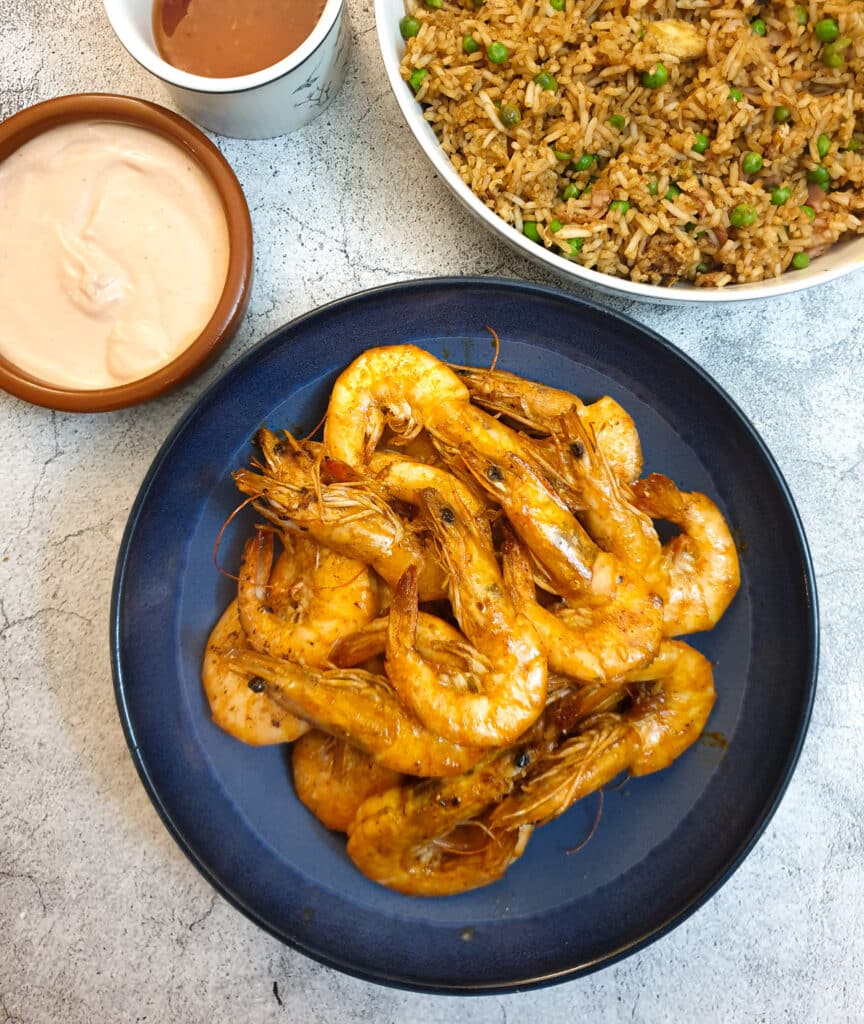 A dish of garlic butter prawns in their shells on a serving dish next to a bowl of fried rice and a pink dipping sauce.