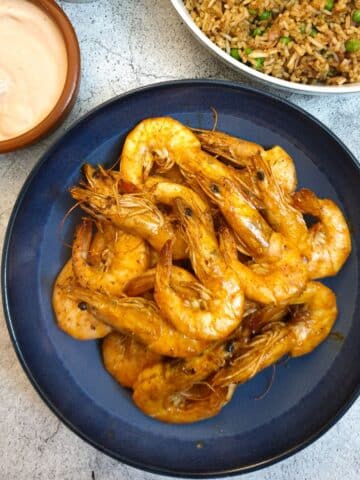 A dish of garlic butter prawns in their shells on a serving dish next to a bowl of fried rice and a pink dipping sauce.