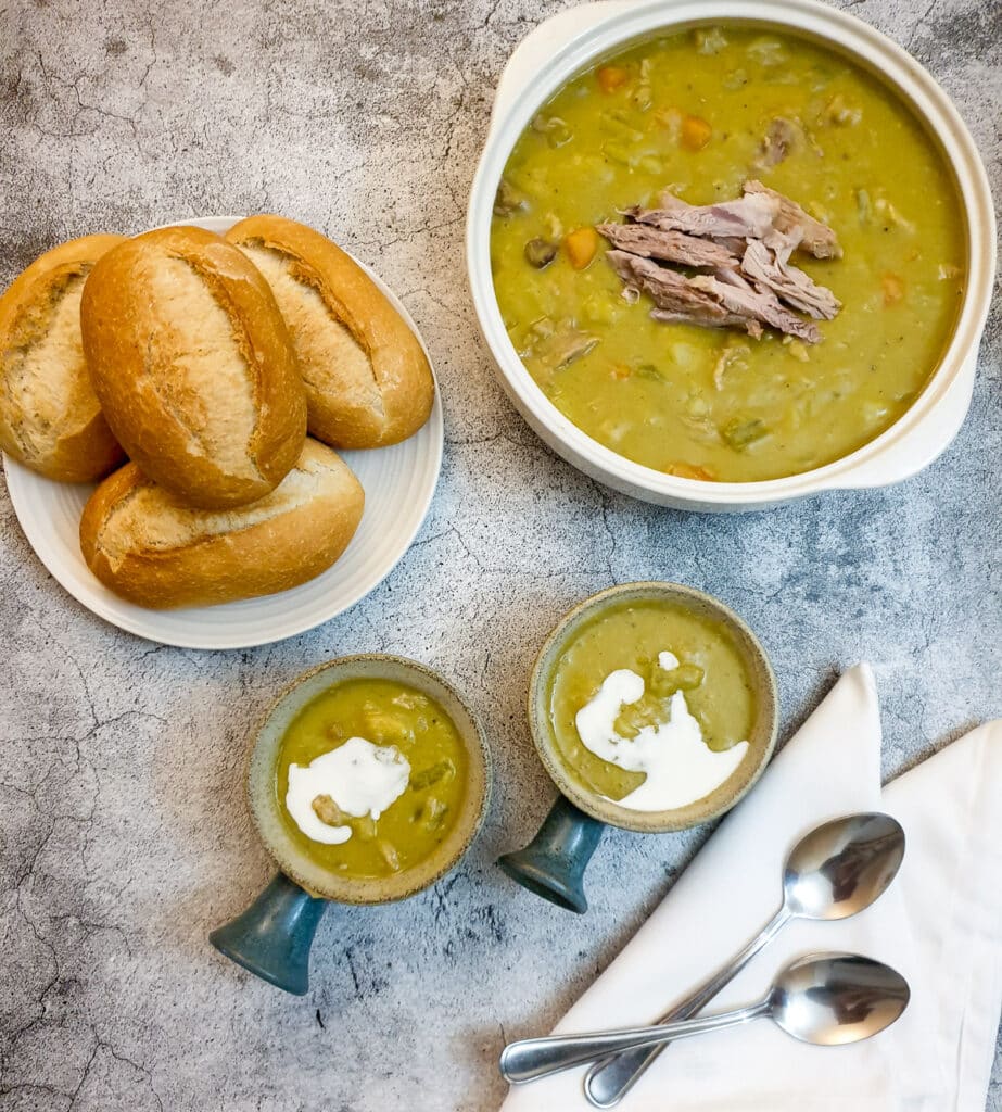 A serving dish of pea and ham soup on a table with crusty bread rolls.