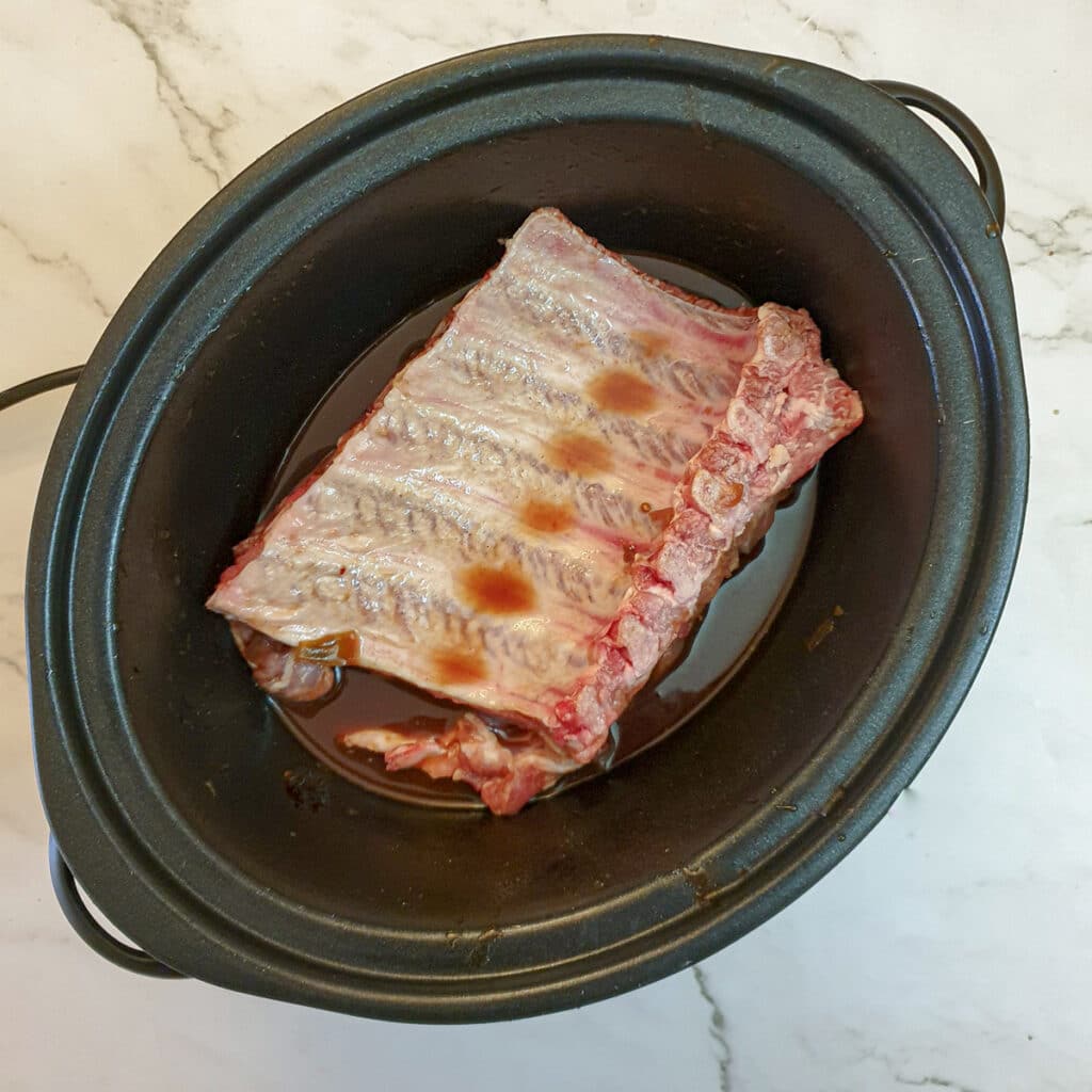 Uncooked pork spare ribs in a slow-cooker, covered with barbeque sauce.