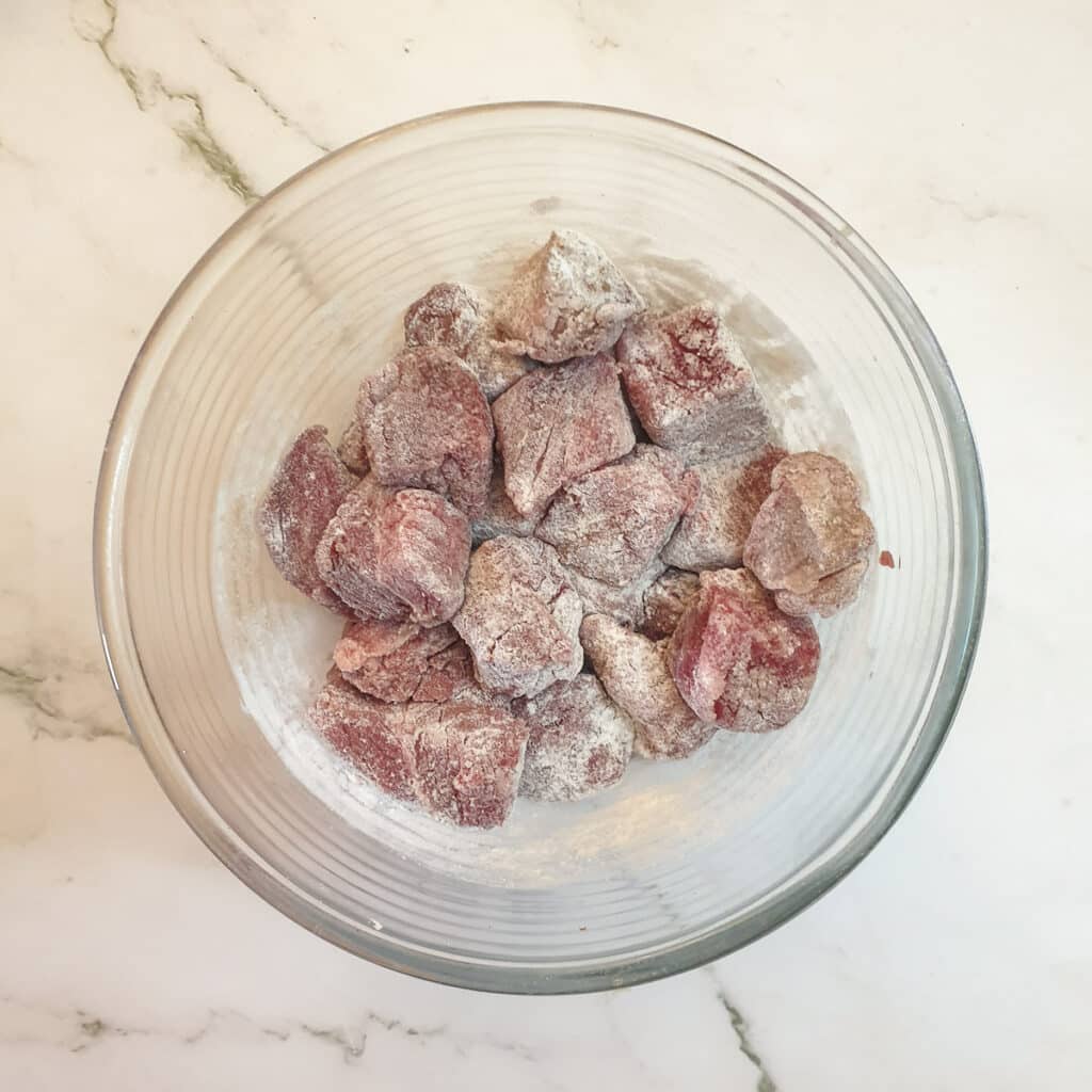 Steak chunks coated in seasoned flour in a glass bowl.