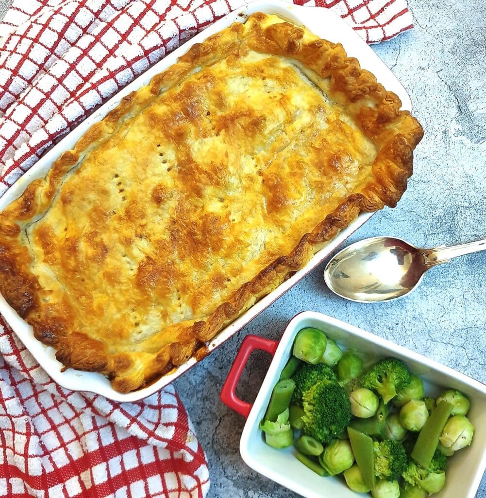 A homemade steak and kidney pie covered with golden crispy puff pastry alongside a dish of brussels sprouts.