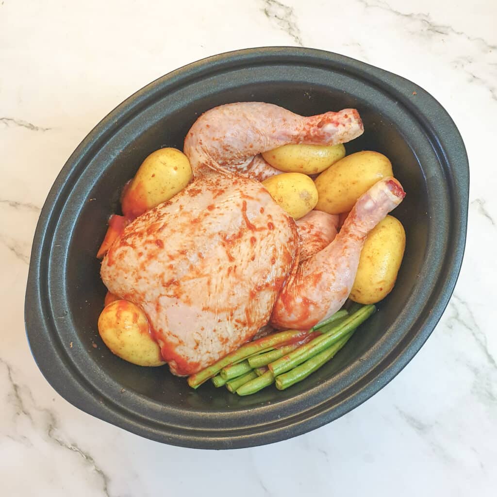 An uncooked chicken on a bed of vegetables in a slow-cooker, covered in gravy.