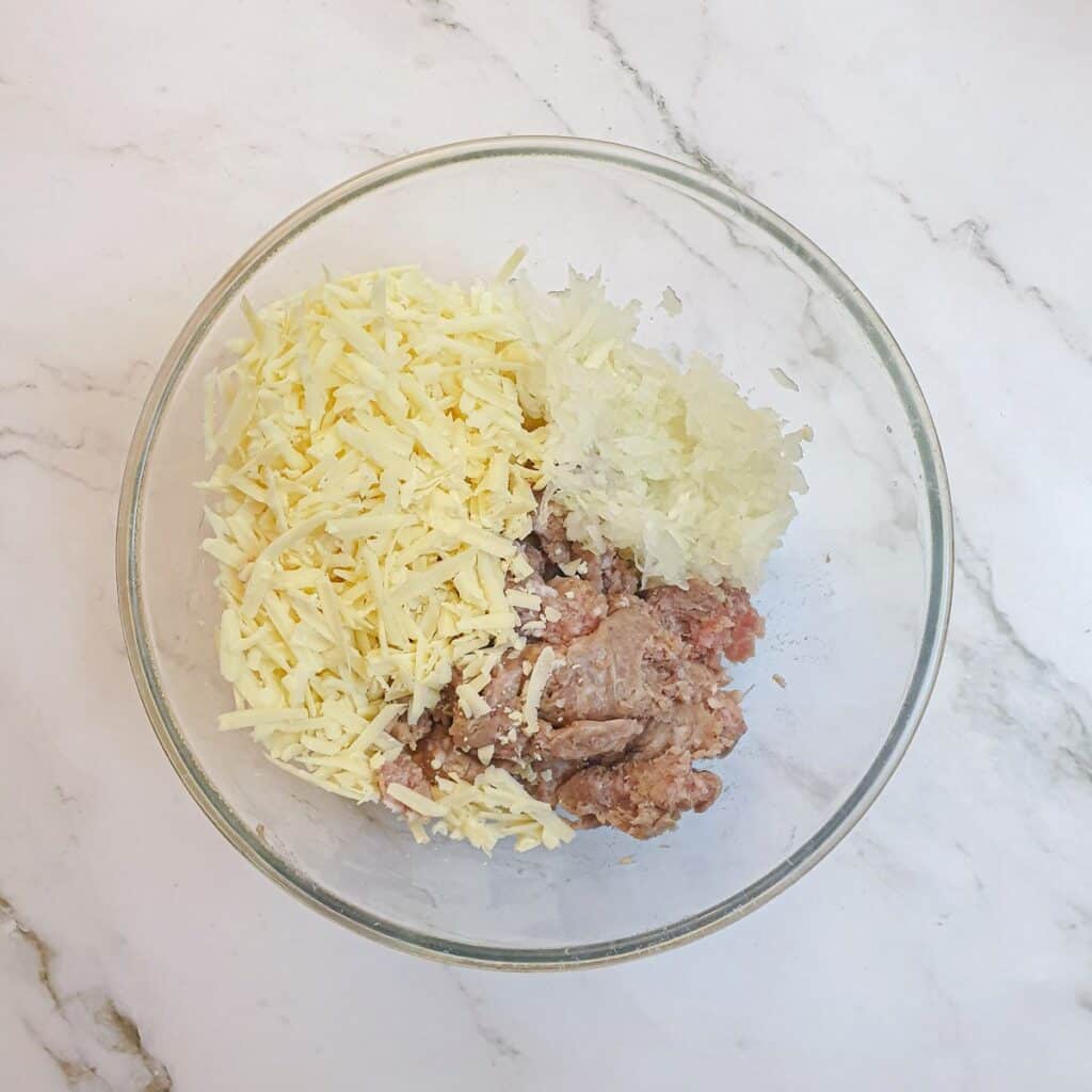 Pork sausage meat, grated onions and grated cheese in a glass mixing bowl.
