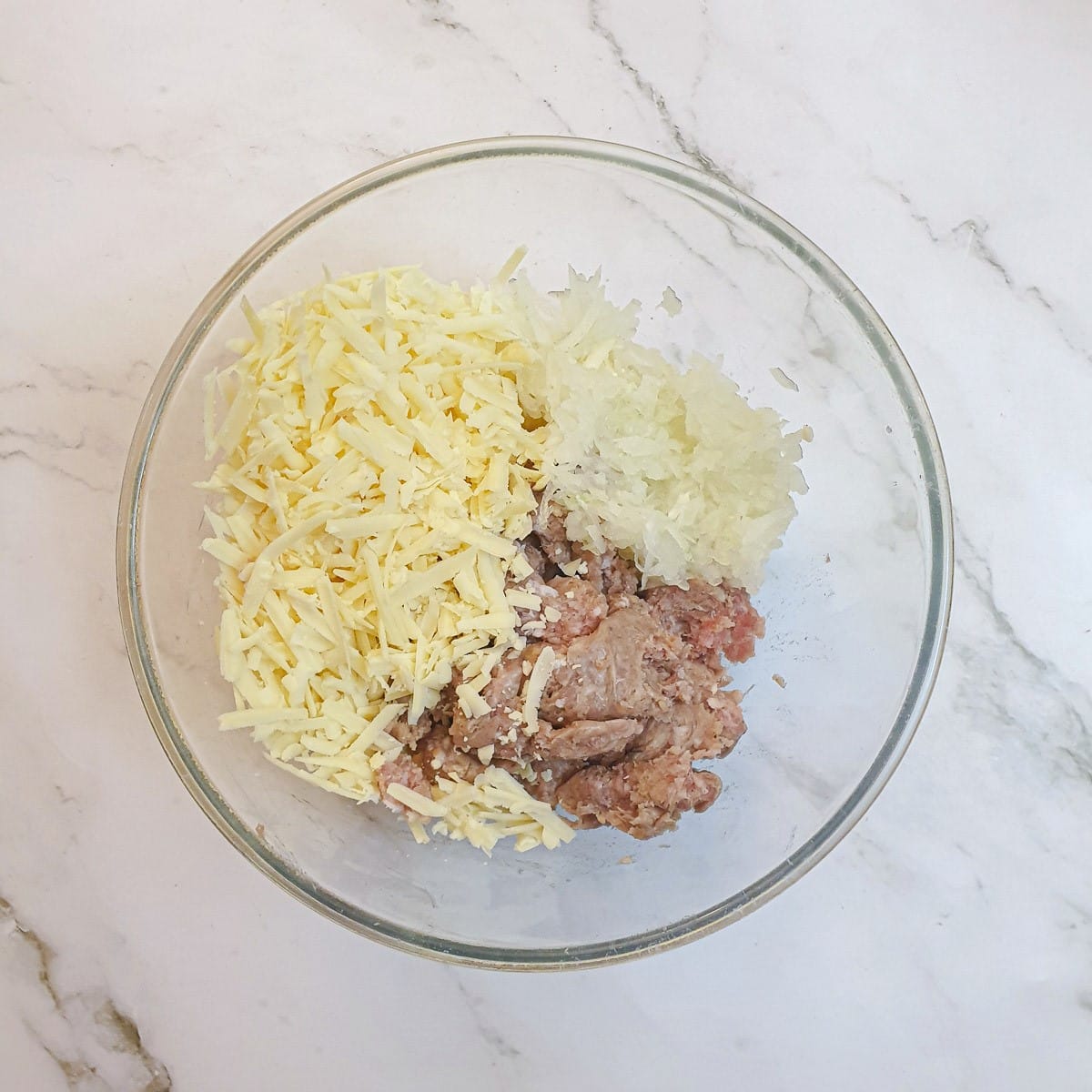 Pork sausage meat, grated onions and grated cheese in a glass mixing bowl.