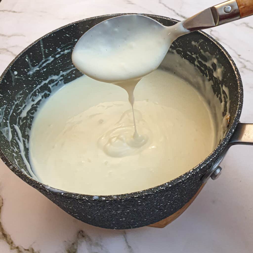 A spoonful of cheese sauce held above a pan.