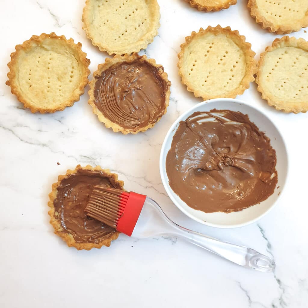 Baked flan cases being brushed with melted chocolate.